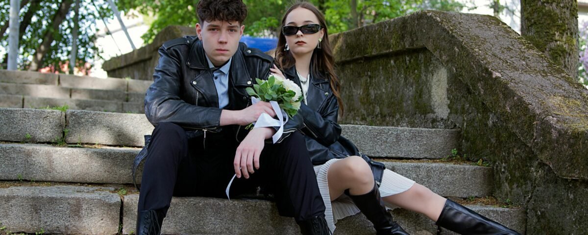Young couple in leather jackets sitting on outdoor steps holding flowers, showcasing modern style.